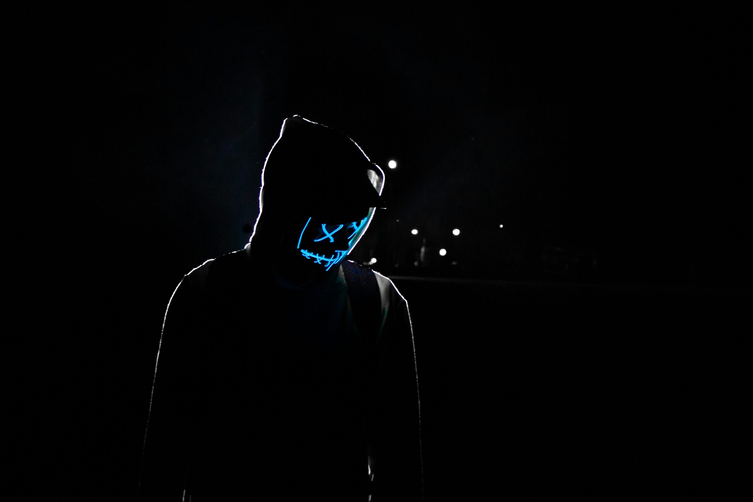 Silhouette of a person in a hoodie wearing a glowing LED mask against a dark background.